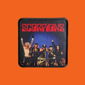 May include: A black square patch with the word 'SCORPIONS' in red lettering. The patch depicts the band Scorpions performing on stage, set against a blue backdrop.