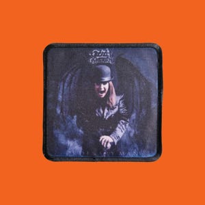 Puede incluir: Parche cuadrado con una imagen oscura de una persona con bombín y chaqueta de cuero con el texto "ORDINARY MAN". El parche tiene un borde negro y está sobre un fondo naranja. La imagen tiene un tema gótico.