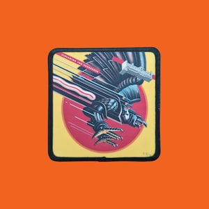 Peut inclure: Écusson carré avec un aigle robotique noir et rouge, avec le texte "Screaming for Vengeance" et "Judas Priest". L'aigle est sur un fond jaune avec un cercle rouge.