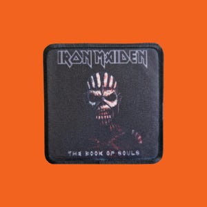 Peut inclure: Écusson carré noir avec le nom du groupe "Iron Maiden" en lettres argentées. L'écusson représente une figure squelettique et le texte "The Book of Souls" sur fond sombre.