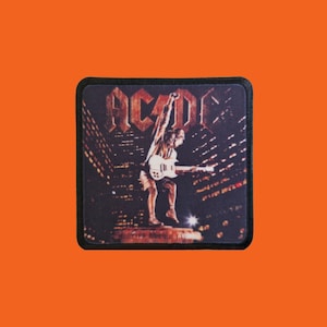 Op de afbeelding: Vierkante patch met het AC/DC-logo en album artwork. De afbeelding toont een gitarist die speelt voor een nachtelijke skyline. De patch heeft een zwarte rand en is geplaatst tegen een oranje achtergrond.