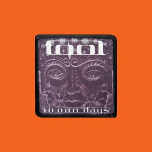 Peut inclure: Un écusson carré à bordure noire. Le nom du groupe "TOOL" est en lettres blanches en haut. En dessous, une image violette d'un visage avec des motifs détaillés. Le texte "10,000 days" est en bas de l'écusson.