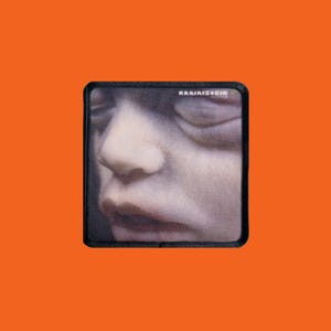 May include: A square patch with a close-up of a human face, featuring a nose, closed eye, and lips. The patch has a black border and the text "KARIMIZ+EIN MUTTER" in white. The background is orange.