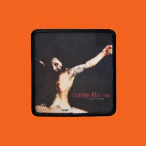 Peut inclure: Écusson carré à bordure noire avec une image stylisée d'une personne avec un bras tendu. Le texte "Marilyn Manson HOLY WOOD" est imprimé en rouge dans le coin inférieur droit.