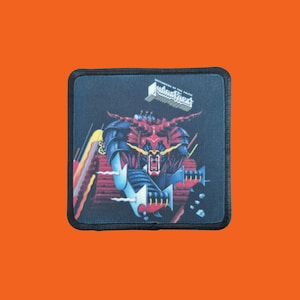 Op de afbeelding: Vierkante patch met een zwarte rand. Het heeft een kleurrijke illustratie van een rode en blauwe robotfiguur met een dreigende uitdrukking. De patch bevat de tekst "Defenders of the Faith".