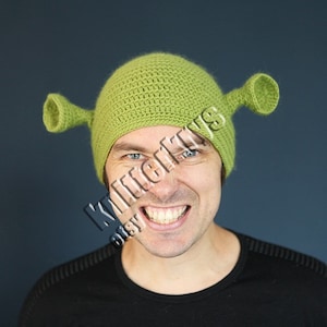Puede incluir: Un gorro de punto verde con dos orejas grandes, que se asemeja al personaje de Shrek.