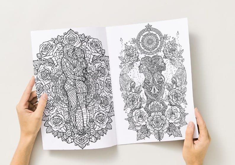 May include: Open coloring book with two detailed black and white illustrations. One depicts a couple embracing within a rose-filled circle. The other shows two faces and a skull surrounded by roses and a mandala.