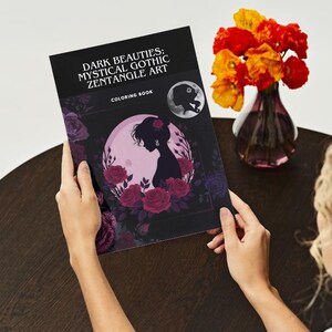 Puede incluir: Un libro para colorear titulado "Dark Beauties: Mystical Gothic Zentangle Art" se sostiene abierto, mostrando la silueta de una mujer rodeada de rosas. Un jarr&oacute;n con flores naranjas y amarillas se encuentra cerca.