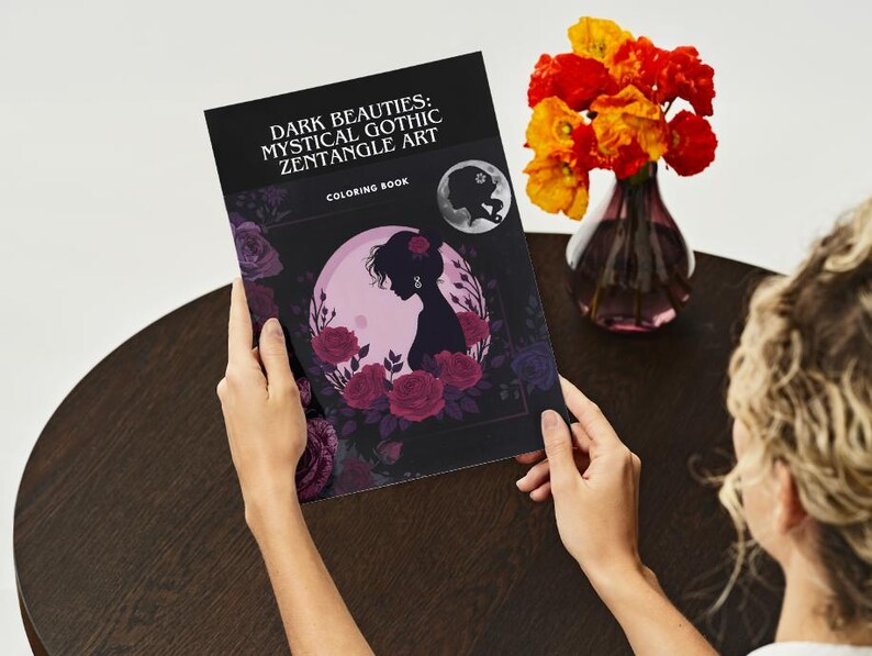 Puede incluir: Un libro para colorear titulado "Dark Beauties: Mystical Gothic Zentangle Art" se sostiene abierto, mostrando la silueta de una mujer rodeada de rosas. Un jarr&oacute;n con flores naranjas y amarillas se encuentra cerca.