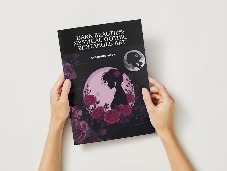 Puede incluir: Un libro para colorear titulado "Dark Beauties: Mystical Gothic Zentangle Art" es sostenido por manos sobre un fondo blanco. La portada presenta una silueta de mujer frente a una luna rosa, rodeada de rosas rojas. Una luna m&aacute;s peque&ntilde;a con una silueta est&aacute; en la esquina superior derecha.