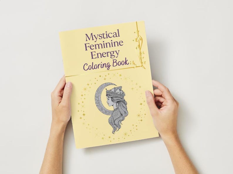 Peut inclure: Un livre de coloriage jaune intitul&eacute; &laquo; Mystical Feminine Energy Coloring Book &raquo; est ouvert. La couverture pr&eacute;sente une illustration du profil d'une femme dans un croissant de lune et des &eacute;toiles. Le livre a des accents dor&eacute;s d&eacute;coratifs.