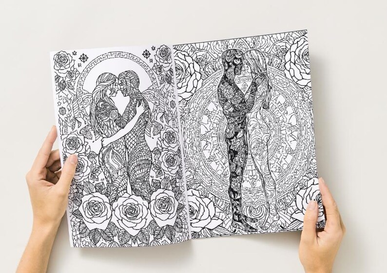 May include: Open coloring book pages featuring intricate black and white illustrations of embracing figures, surrounded by floral and geometric patterns. The pages are held open, showcasing the detailed artwork.