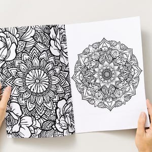 May include: Two open pages of a coloring book featuring intricate black and white floral and mandala designs. The left page displays a floral pattern, while the right page showcases a detailed mandala. The pages are held open by hands.