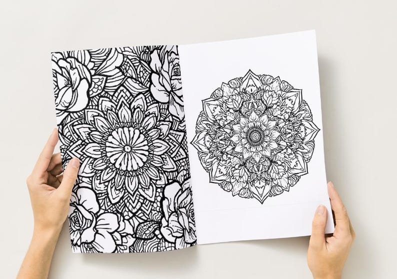 May include: Two open pages of a coloring book featuring intricate black and white floral and mandala designs. The left page displays a floral pattern, while the right page showcases a detailed mandala. The pages are held open by hands.