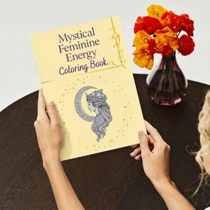 Peut inclure: Un livre de coloriage jaune intitul&eacute; "Mystical Feminine Energy Coloring Book" est ouvert, r&eacute;v&eacute;lant une illustration du profil d'une femme dans un croissant de lune. Un vase de fleurs orange et rouges est &agrave; proximit&eacute;.