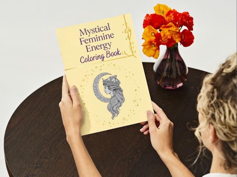 Peut inclure: Un livre de coloriage jaune intitul&eacute; "Mystical Feminine Energy Coloring Book" est ouvert, r&eacute;v&eacute;lant une illustration du profil d'une femme dans un croissant de lune. Un vase de fleurs orange et rouges est &agrave; proximit&eacute;.