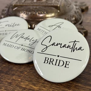 May include: A collection of light green round buttons with black text. One button reads "Samantha BRIDE" in a script font. Other buttons have names and titles like "Maid of Honor". The buttons are on a dark wooden surface.