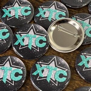 Collection of team buttons with the logo XTC on a wooden surface. These round custom button badges are great for business branding, events, school activities, campaigns and giveaways.