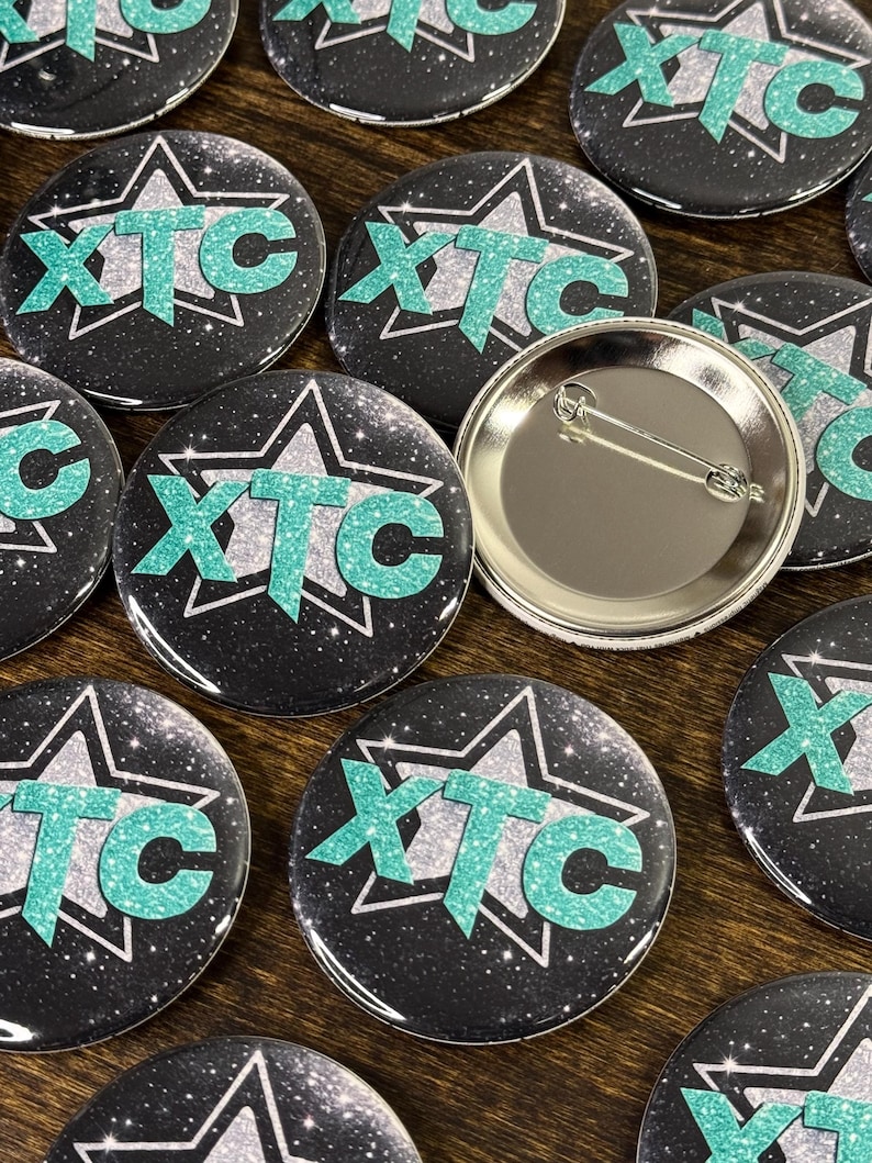 Collection of team buttons with the logo XTC on a wooden surface. These round custom button badges are great for business branding, events, school activities, campaigns and giveaways.