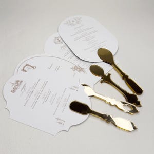 Peut inclure: Faire-part de mariage blancs avec texte doré et éléments décoratifs, accompagnés de cuillères dorées. Les invitations présentent les détails de l'événement et un programme. Les cuillères sont disposées près des invitations.