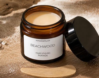 May include: An amber glass jar of Wanderlox Beachwood hair styling pomade with a black lid, containing a light-colored cream. A small wooden applicator tool lies in front of the open jar, which features a white label with black text.