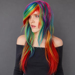Könnte beinhalten: Eine lange, gestufte Perücke mit einer lebendigen Regenbogen-Farbpalette mit Strähnen in Rot, Orange, Gelb, Grün, Blau und Violett. Das Haar ist mit einem seitlichen Pony gestylt und fällt glatt und geschmeidig über die Schultern.