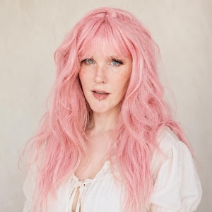 May include: A person with long, wavy, light pink hair and bangs. The person is wearing a white, long-sleeved top with a lace-up neckline. The background is a neutral, off-white color.
