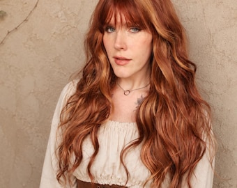 Puede incluir: Una persona con largo cabello castaño rojizo ondulado con flequillo, que lleva una blusa color crema y un corsé marrón. Se ve un delicado collar de plata. El fondo es una pared texturizada de color marrón claro con una planta trepadora.