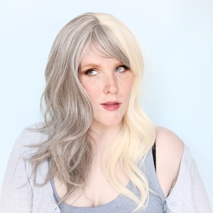 May include: A woman with half of her hair dyed a light blonde color and the other half a silver grey color. She is wearing a light grey cardigan and a grey tank top.