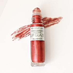 May include: A bottle of Miss Violet Lace Pomegranate Sugar Lip Glaze, with a red, glittery finish. The product is displayed on a white surface with a matching lip glaze swatch. The bottle is labeled with the product name and volume.