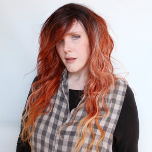 May include: A woman with long, wavy hair that is dyed a reddish-orange color with blonde highlights. She is wearing a black shirt and a plaid vest.