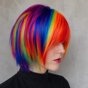 May include: A person with a striking rainbow-coloured hairstyle, showcasing red, orange, yellow, green, blue, and purple hues. The hair is cut short and layered. They are wearing a black turtleneck top.