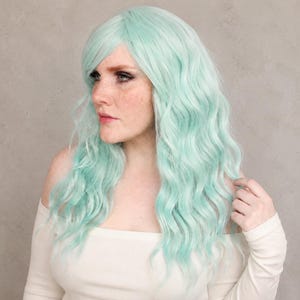 K&ouml;nnte beinhalten: Eine lange, gewellte Per&uuml;cke in blassem Mintgr&uuml;n. Die Per&uuml;cke hat einen seitlich gescheitelten Pony und lockere, flie&szlig;ende Wellen, die &uuml;ber die Schultern fallen. Das Haar hat eine weiche, matte Textur und einen gleichm&auml;&szlig;igen Farbton.