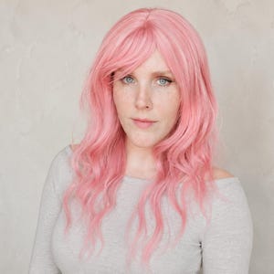 Könnte beinhalten: Eine lange, gewellte, pastellrosa Perücke mit seitlich gescheiteltem Pony und gestuften Strähnen, die über die Schultern fallen. Das Haar hat eine weiche, matte Textur und eine gleichmäßige hellrosa Farbe über die gesamte Haarlänge.