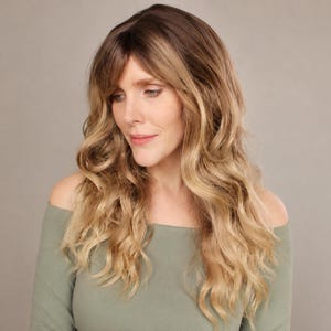 May include: A woman with long, wavy blonde hair and a dark root. The hair cascades over the shoulders. She is wearing a sage green off-the-shoulder top. The background is a solid, neutral color.