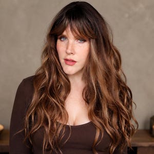 May include: A woman with long, wavy brown hair and bangs, wearing a dark brown long-sleeved top. She has fair skin, blue eyes, and is looking directly at the camera. Candles and a decorative lantern are on a wooden surface in the background.