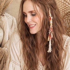 May include: A woman with long, wavy auburn hair, adorned with a decorative hair accessory featuring shells, beads, and fabric. She wears a cream-colored blouse with lace detailing, and smiles. The background includes a wicker chair and a vase with dried wheat.