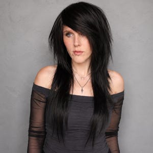 May include: A person with long, layered black hair and blue eyes, wearing a dark gray off-the-shoulder top and a silver pendant necklace. The background is a solid gray.