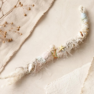 May include: A handcrafted, bohemian-style decorative piece. It features a wrapped, off-white base adorned with seashells, a small starfish, and delicate strands. The design incorporates light blue, pink, and green accents, creating a coastal aesthetic.