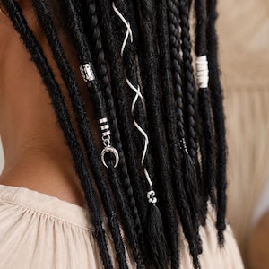 May include: Close-up of black dreadlocks adorned with silver and white hair accessories. The dreadlocks are styled with decorative beads and charms, including a crescent moon pendant. The hair is styled with white and silver spiral accents.