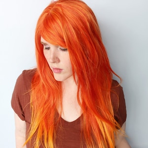 May include: A woman with long, bright orange hair with yellow highlights. The hair is styled in loose waves and falls past her shoulders. She is wearing a brown t-shirt.