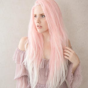 May include: A long, layered wig with a gradient of pink to white. The wig is styled with long, flowing strands. The person is wearing a mauve off-the-shoulder dress with ruffled sleeves. The background is a neutral, textured wall.