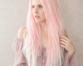 Op de afbeelding: Een lange, gelaagde pruik met een verloop van roze naar wit. De pruik is gestyled met lange, vloeiende lokken. De persoon draagt een mauvekleurige off-the-shoulder jurk met ruchesmouwen. De achtergrond is een neutrale, getextureerde muur.