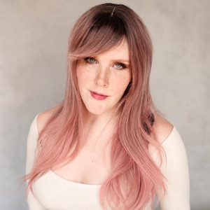 May include: A woman with long, wavy, pink hair with dark roots and bangs. She is wearing a white off-the-shoulder top. The background is a solid, light gray color.