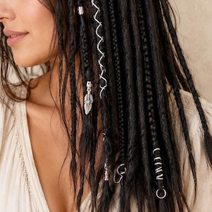 May include: Close-up of black braided hair adorned with silver hair accessories. The hair features decorative metal spirals, rings, and charms, including a feather pendant with a turquoise stone. The person is wearing a cream-colored top.