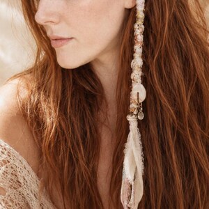 May include: A woman with long auburn hair, adorned with a decorative hair accessory. The accessory is a strand of white fabric, shells, and beads. She wears a cream-colored, open-shoulder lace top.