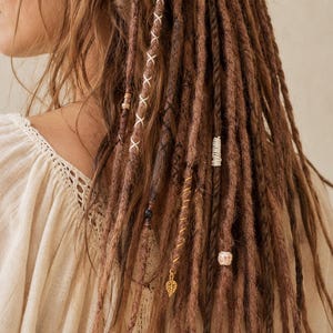 May include: Close-up of brown dreadlocks with decorative beads and charms. The hair is styled with white thread accents and a gold leaf charm. The person is wearing a cream-colored, long-sleeved top with a lace neckline.