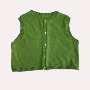 May include: A sleeveless, button-down sweater vest in a vibrant green. The vest features a crew neck and is fastened with small, white buttons down the centre. The knitted texture adds visual interest to this casual garment.