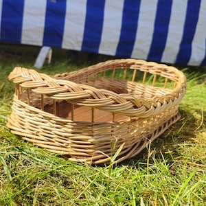 Könnte beinhalten: Ein geflochtener Weidenkorb mit geflochtenem Rand liegt auf grünem Gras. Der Korb hat eine rechteckige Form mit gewölbter Oberseite. Ein blau-weiß gestreifter Stoff bildet den Hintergrund.