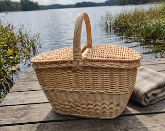 May include: A light brown woven wicker picnic basket featuring a sturdy central handle and two hinged lids that open from the middle. The basket has a rounded rectangular shape with a tight, uniform weave pattern throughout the body and lids.